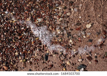 Close-up picture of sandy beach sand with small colorful stones rocks and foamy waves of sea water. Natural texture tropical background.