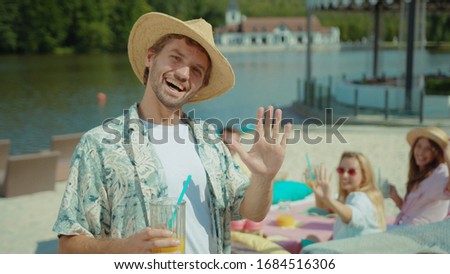 Portrait of happy young stylish man drinking juice cocktail staying at lake beach. Excited friends enjoying drinks and food on beach calling him to join summer party.
