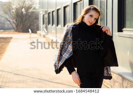 Portrait of a stylish young girl with long hair with a smile in the city against the background of the wall of a business building with windows. Photo taken in sunny weather outdoors in spring.