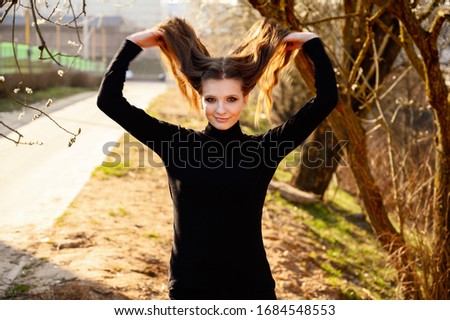 Portrait of a pretty young girl with long hair with a smile on the background of road trees in the park. Photo in sunny weather outdoors in spring.