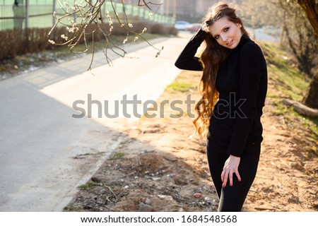 Portrait of a pretty young girl with long hair with a smile on the background of road trees in the park. Photo in sunny weather outdoors in spring.