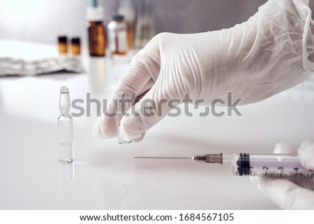 Female hands in white medical gloves, right hand picking up one of two ampoules with medicine from white acrylic table, left hand holding a syringe. Ampoules, glass jars, blisters on background