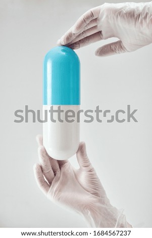 Female hands in white medical gloves holding a large plastic blue with white pill vertically against light background
