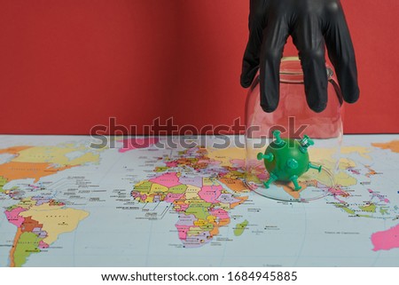 Conceptual photo of a hand protected with a black glove catching a green virus with a transparent glass on a world political map on a red background. The virus is on asia.