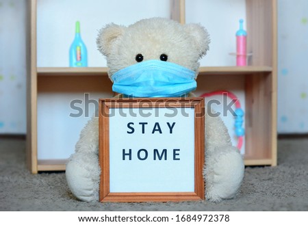 Stay at home, virus epidemic warning, quarantine, no kindergarten: teddy bear in medical face mask sits near children playhouse and holding hands poster, picture frame with the words «Stay home»