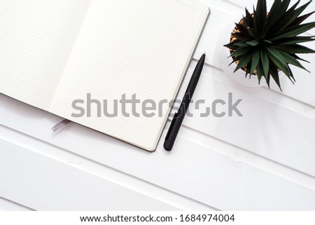 A blank white notebook on a white wooden background next to a home flower in a pot. Light pattern