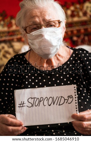 An elderly woman in a medical mask. holding a tablet with an inscription #stopcovid-19