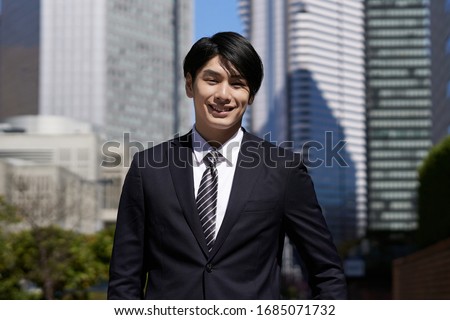 A smiling Japanese male businessman stands in an office district.