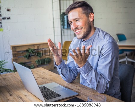 happy smiling remote online working man having his hand up with laptop and notebook in casual outfit with sitting in an coworking or home office at a work desk explaining something
