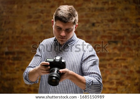 Front view of a Caucasian male photographer taking pictures with a digital SLR camera in a photography studio and checking the images on the camera display with brick wall in the background.