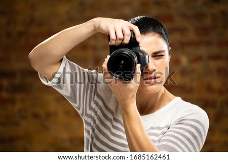 Front view of a Caucasian female photographer with long dark hair taking pictures with a digital SLR camera in a photography studio with brick wall in the background.
