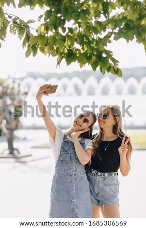 two young pretty girls on a walk in the park taking pictures of themselves on the phone. selfies on a sunny summer day, joy and friendships.