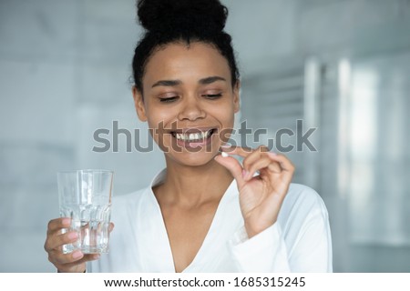 Head shot african woman holds pill and glass of water. Taking minerals for female skin and health, boost metabolism, vitamin A C deficiency prevention treatment, healthcare, skincare bodycare concept Royalty-Free Stock Photo #1685315245