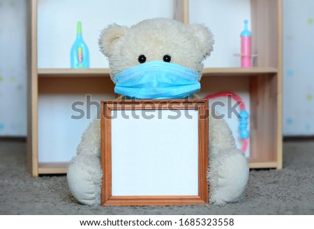 Copy spaсe in holding hands - for theme quarantine, stay at home, coronavirus.
Teddy bear in medical face mask sits near children playhouse and holds empty picture frame with place for inscription.