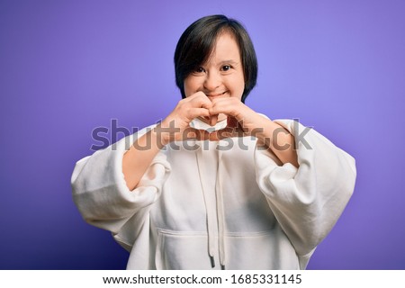 Young down syndrome sports woman wearing fitness sweatshirt over purple background smiling in love showing heart symbol and shape with hands. Romantic concept.
