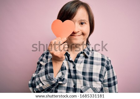 Young down syndrome romantic woman holding red heart paper shape over pink background with a happy face standing and smiling with a confident smile showing teeth