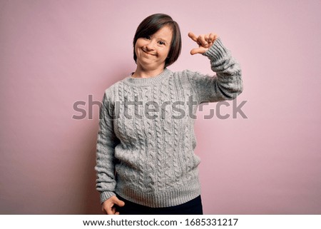Young down syndrome woman wearing casual sweater over isolated background smiling and confident gesturing with hand doing small size sign with fingers looking and the camera. Measure concept.