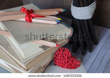 Books, colored pencils between pages, pencils tied with a red satin ribbon on a stack, a bunch of wooden twigs standing next to them
