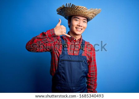 Young handsome chinese farmer man wearing apron and straw hat over blue background smiling doing phone gesture with hand and fingers like talking on the telephone. Communicating concepts.