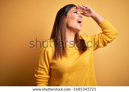 Young beautiful woman wearing casual sweater over yellow isolated background very happy and smiling looking far away with hand over head. Searching concept.