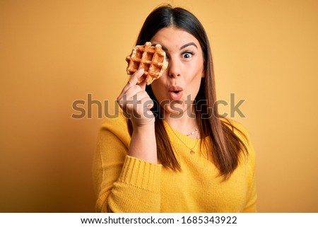 Young beautiful woman eating sweet waffle pastry over yellow background scared in shock with a surprise face, afraid and excited with fear expression