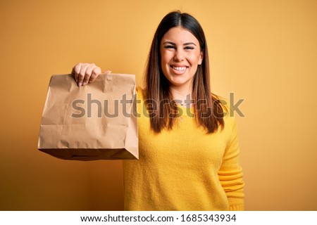Young beautiful woman holding take away paper bag from delivery over yellow background with a happy face standing and smiling with a confident smile showing teeth