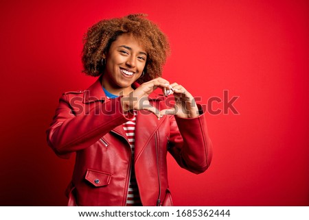 Young beautiful African American afro woman with curly hair wearing casual red jacket smiling in love doing heart symbol shape with hands. Romantic concept.