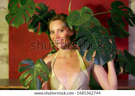 Woman in a trendy modern olive-colored dress next to the evergreen monstera in a white pot on a red background.