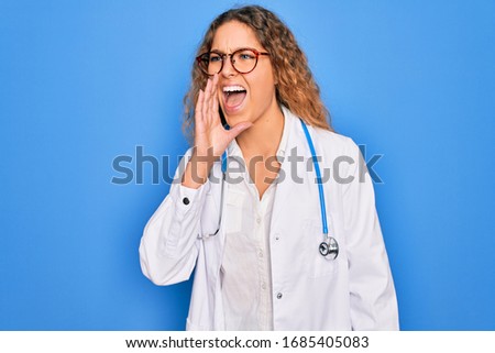 Young beautiful blonde doctor woman with blue eyes wearing coat and stethoscope shouting and screaming loud to side with hand on mouth. Communication concept.