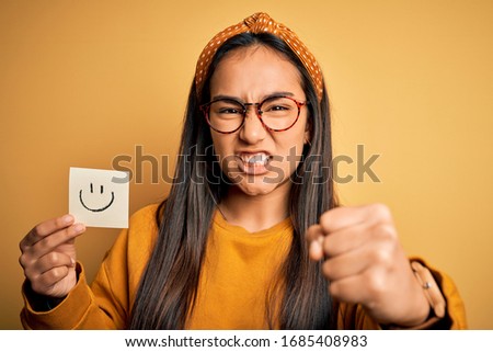 Asian woman wearing glasses holding reminder with smile emoji draw over yellow background annoyed and frustrated shouting with anger, crazy and yelling with raised hand, anger concept