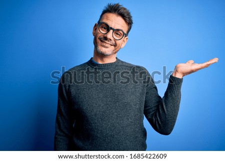 Young handsome man wearing casual sweater and glasses standing over blue background smiling cheerful presenting and pointing with palm of hand looking at the camera.