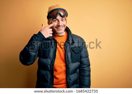 Young handsome skier man skiing wearing snow sportswear using ski goggles Pointing with hand finger to face and nose, smiling cheerful. Beauty concept