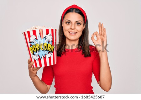 Young beautiful brunette woman eating pack of popcorns snack over isolated white background doing ok sign with fingers, excellent symbol
