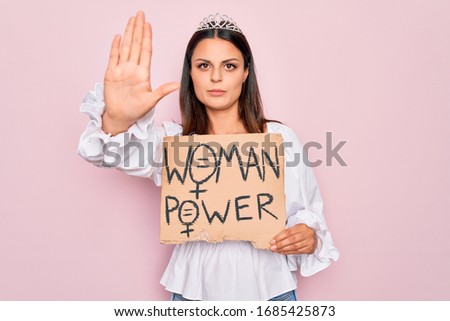 Girl wearing princess tiara asking for women rights holding banner with woman power message with open hand doing stop sign with serious and confident expression, defense gesture