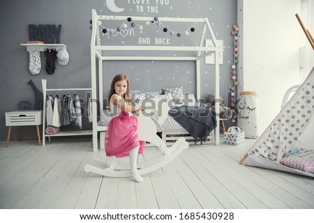 Little, beautiful, charming girl child sitting on a wooden horse in a bright room with a modern interior, pillow decor