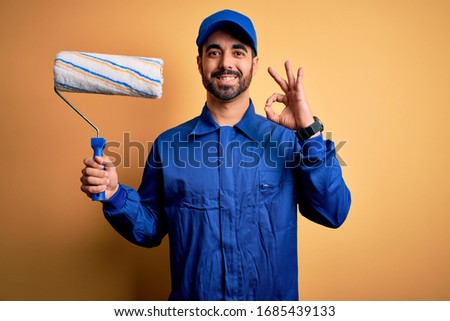 Young handsome painter man with beard wearing blue uniform and cap painting using roller doing ok sign with fingers, excellent symbol