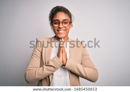 Beautiful african american businesswoman wearing jacket and glasses over white background praying with hands together asking for forgiveness smiling confident.