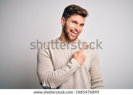 Young handsome blond man with beard and blue eyes wearing casual sweater cheerful with a smile of face pointing with hand and finger up to the side with happy and natural expression on face