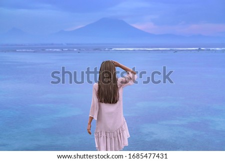 A beautiful girl with long brunette hair in a pink dress stands on top of the rock above the sea and looks at the mountains in the distance. Mystical picture at sunrise. Agung volcano at dawn. Sunrise