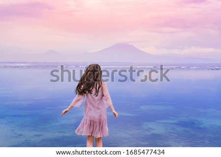 Adventurous beautiful girl with long brunette hair in a pink dress stands on top of the rock above the ocean and looks at the mountains in the distance. Mystical picture at sunset. Agung volcano at