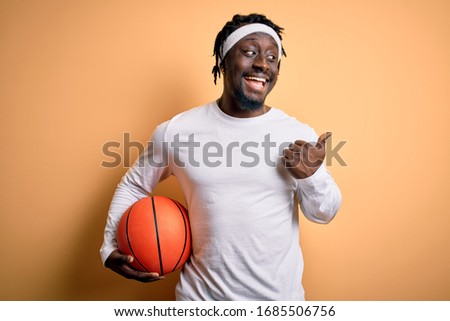 Young african american sportsman doing sport holding basketball ball over yellow background pointing and showing with thumb up to the side with happy face smiling