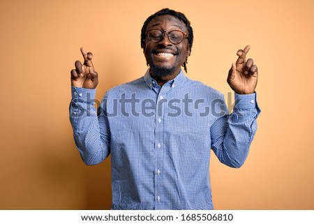 Young handsome african american man wearing shirt and glasses over yellow background gesturing finger crossed smiling with hope and eyes closed. Luck and superstitious concept.