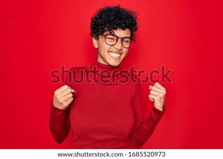 Young beautiful african american afro woman wearing turtleneck sweater and glasses very happy and excited doing winner gesture with arms raised, smiling and screaming for success. Celebration concept.