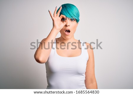 Young beautiful woman with blue fashion hair wearing casual t-shirt over white background doing ok gesture shocked with surprised face, eye looking through fingers. Unbelieving expression.