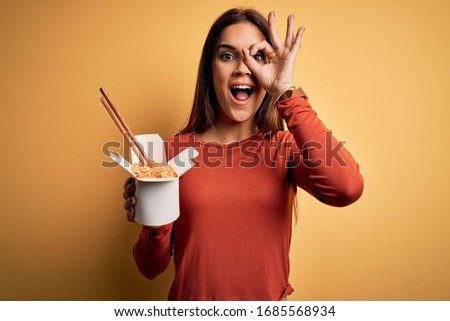 Young beautiful brunette woman eating takeaway noodles using chopsticks with happy face smiling doing ok sign with hand on eye looking through fingers