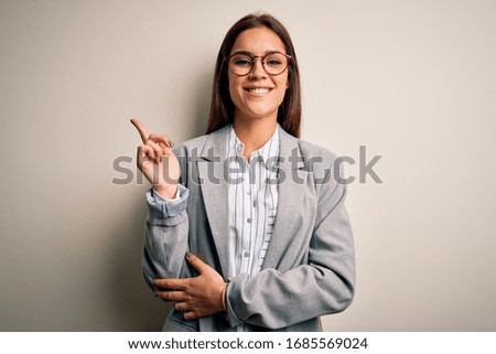 Young beautiful brunette businesswoman wearing jacket and glasses over white background with a big smile on face, pointing with hand and finger to the side looking at the camera.