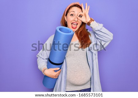 Young beautiful redhead pregnant woman expecting baby holding mat to do prenatal exercise with happy face smiling doing ok sign with hand on eye looking through fingers