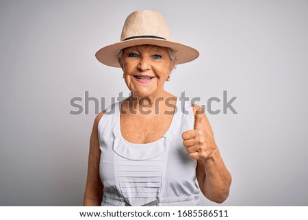 Senior beautiful grey-haired woman on vacation wearing casual summer dress and hat doing happy thumbs up gesture with hand. Approving expression looking at the camera showing success.