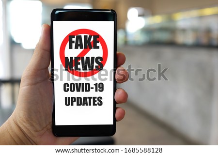 Close up adult hand holding a smartphone with Stop Fake News of Covid-19 Updates text and symbol on display screen, blurred cafe background. Royalty-Free Stock Photo #1685588128