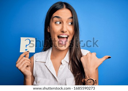 Young beautiful brunette woman holding paper with question mark symbol message pointing and showing with thumb up to the side with happy face smiling